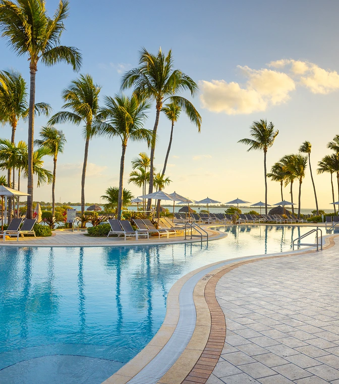 Tropical resort pools and saltwater lagoon lined with palm trees and lounge chairs at sunset.