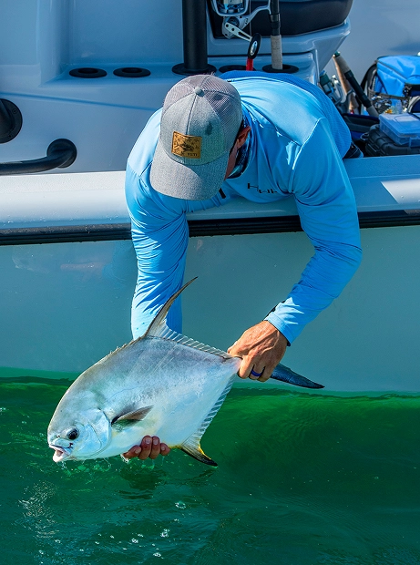 A person on a boat leaning over to carefully release a silvery permit fish into clear turquoise water.