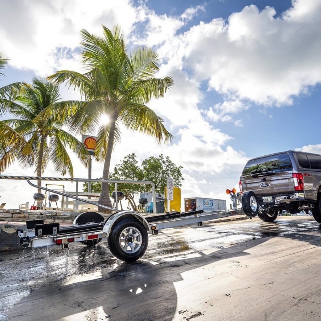 Pickup truck towing a boat trailer near palm trees at a waterfront launch on a sunny day.