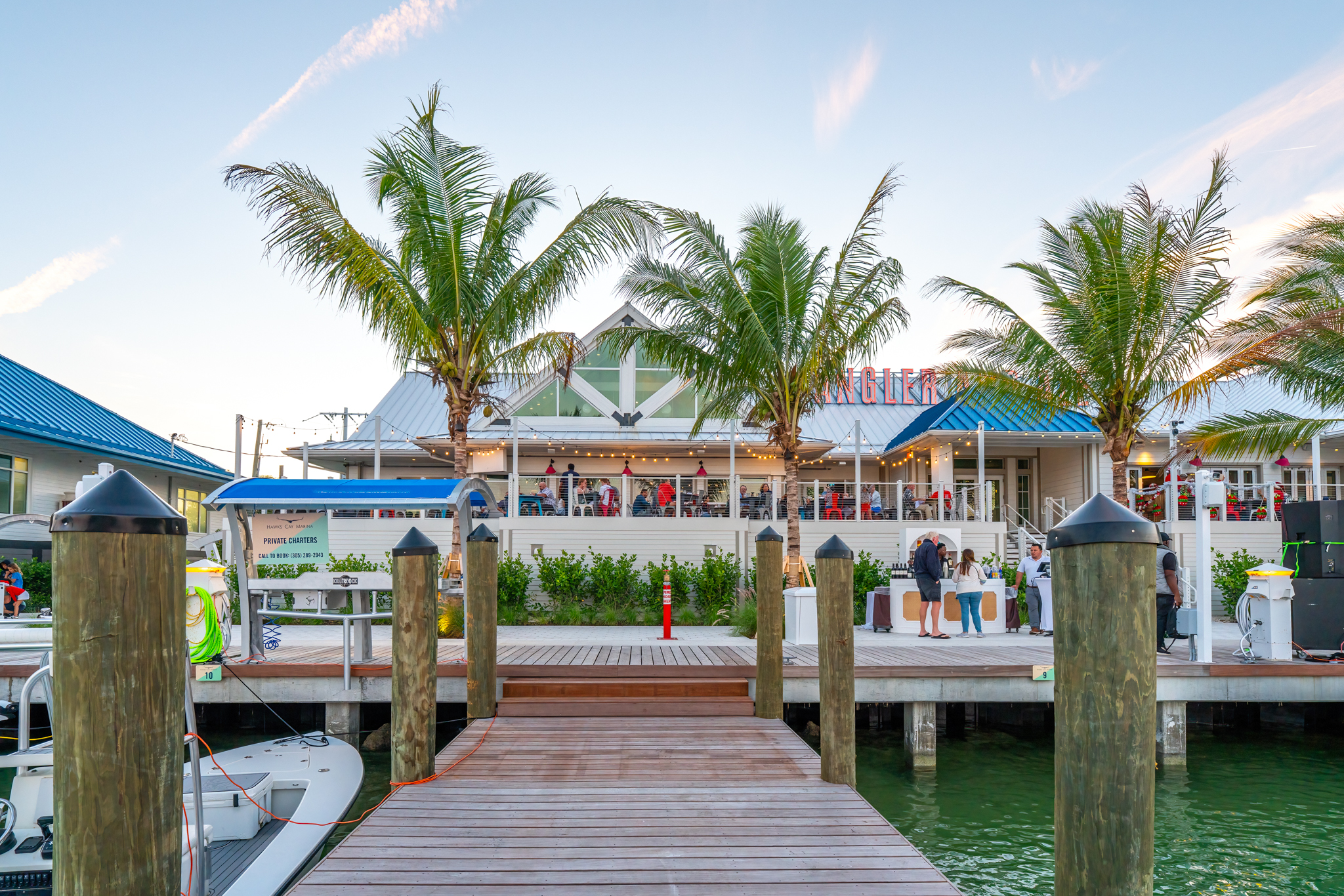 Waterfront restaurant with palm trees, outdoor seating, and a wooden dock overlooking green marina water at sunset.
