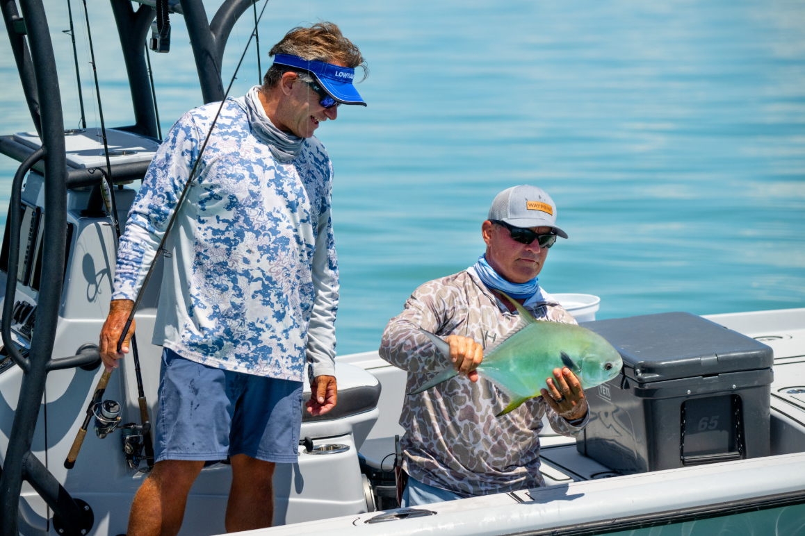 Two men fishing on a boat, one holding a green fish over the deck with calm blue water in the background.