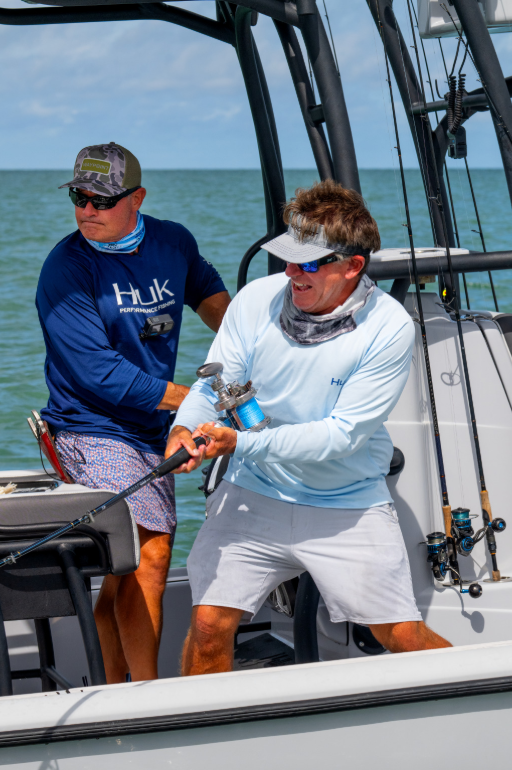 Two men on a fishing boat reeling in a fish together, wearing sun-protective shirts with ocean in the background.