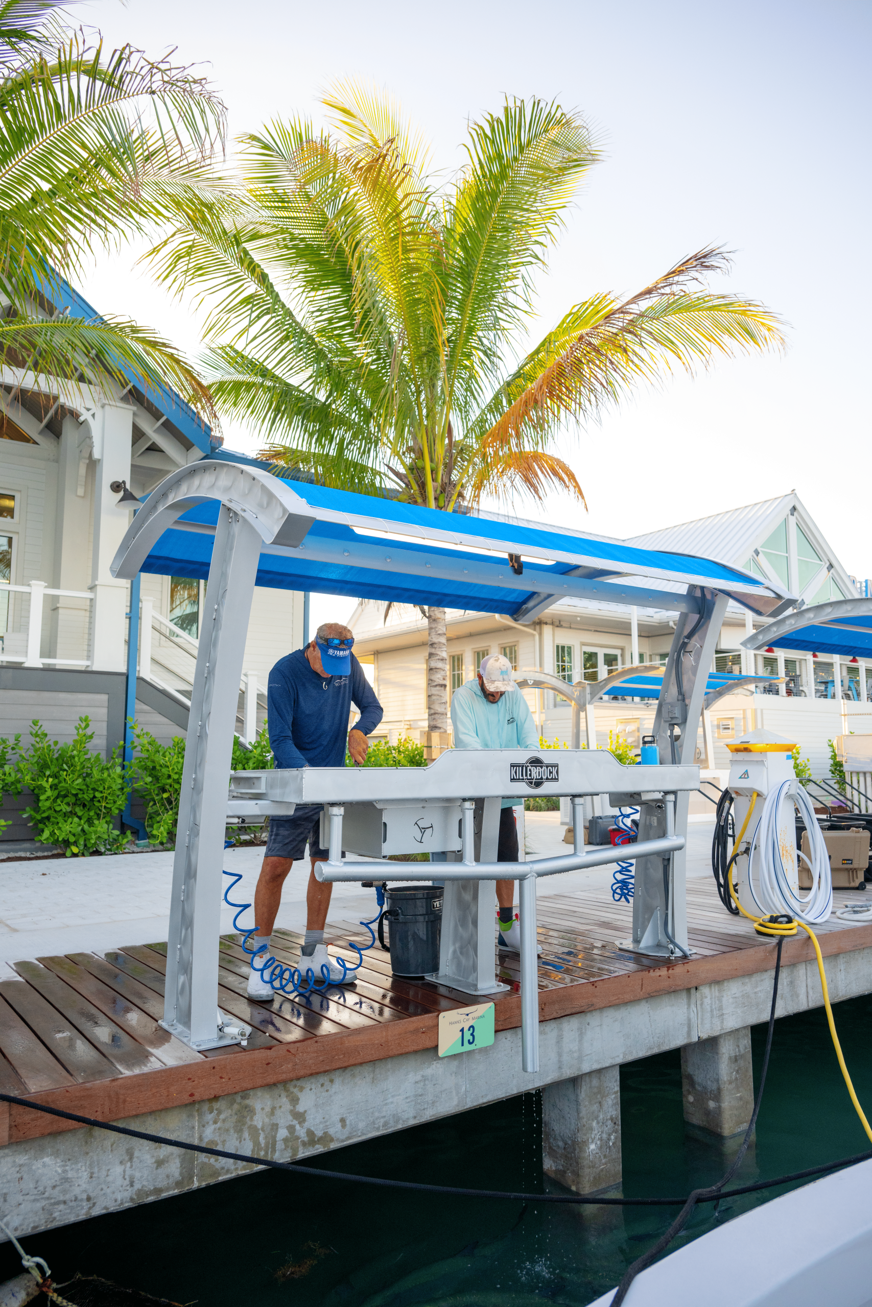 Two people cleaning fish at an outdoor dock station with palm trees and waterfront buildings in the background.
