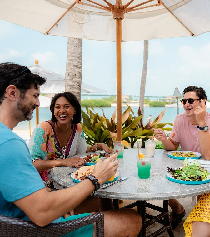 Friends laughing and enjoying tropical drinks and lunch at a shaded waterfront table by a resort lagoon.