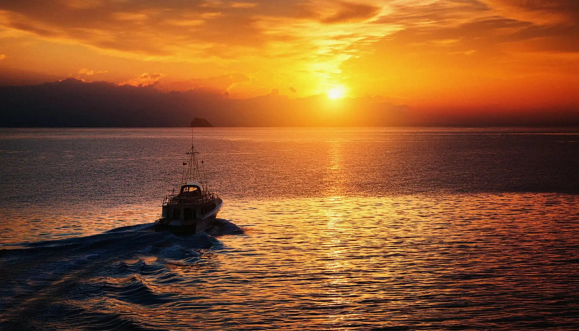 A boat cruising through calm ocean waters toward a vibrant orange and yellow sunset on the horizon.