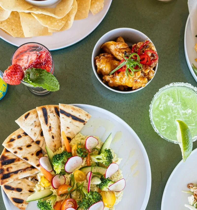 Overhead view of tropical cocktails and appetizers, including hummus and wings, at Pilar Bar.