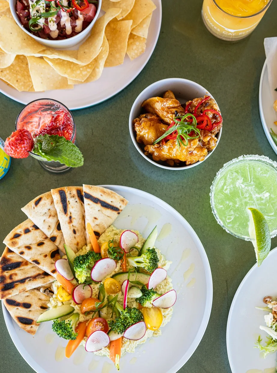 Overhead view of colorful shared dishes with flatbread, vegetables, chips, small plates, and cocktails on a green table.