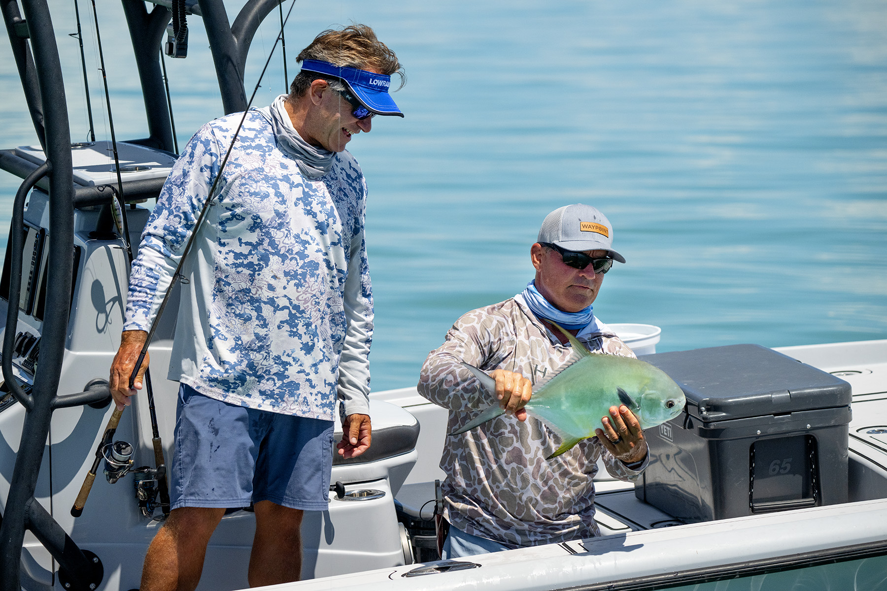 Two people on a fishing boat holding a rod and a freshly caught permit fish, with sun gear and YETI cooler.