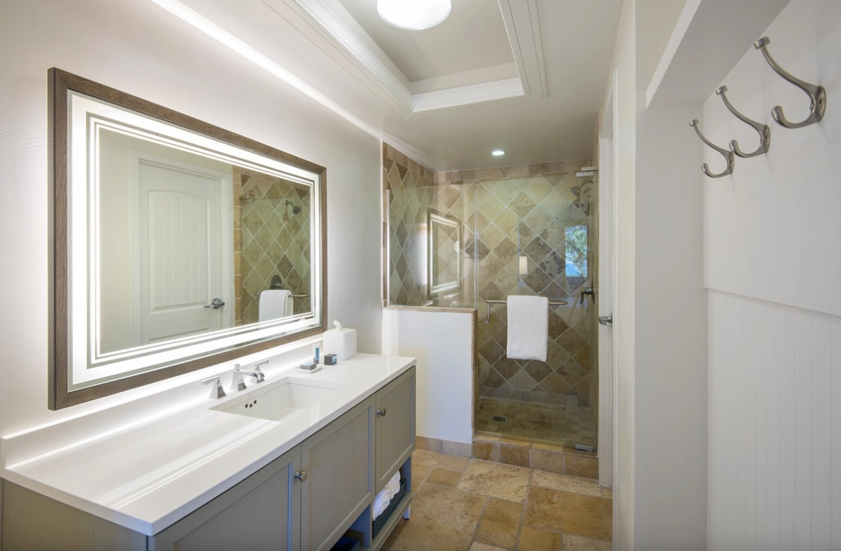 Modern bathroom with a lighted mirror, grey vanity, and a glass walk-in shower with tan stone tile.