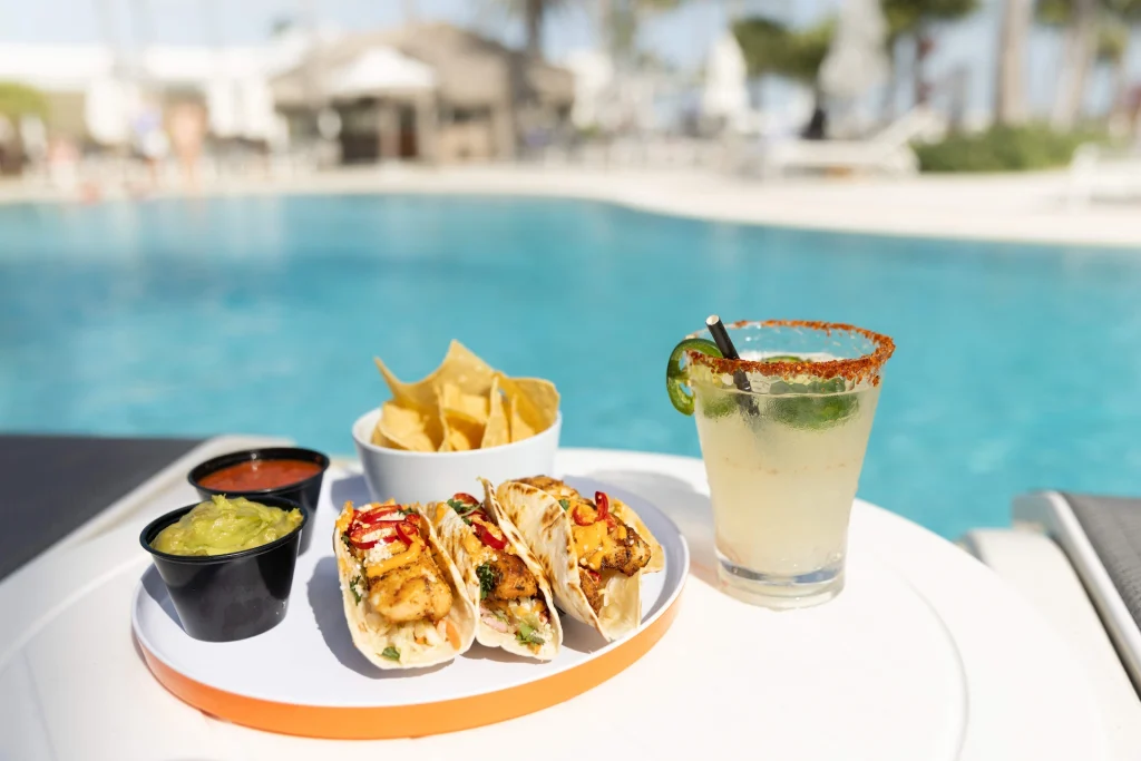 Fish tacos with chips, salsa, guacamole, and a margarita on a table by a bright blue resort pool.
