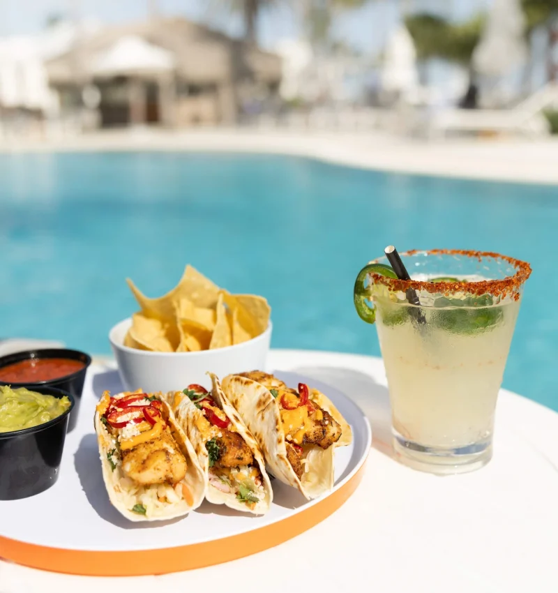Fish tacos with chips, salsa, guacamole, and a margarita on a table by a bright blue resort pool.