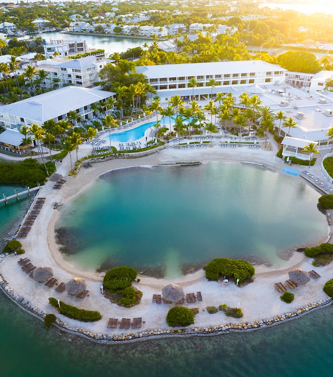 Aerial view of a tropical resort with a saltwater lagoon, sandy beach, swimming pools, and palm trees at sunset.