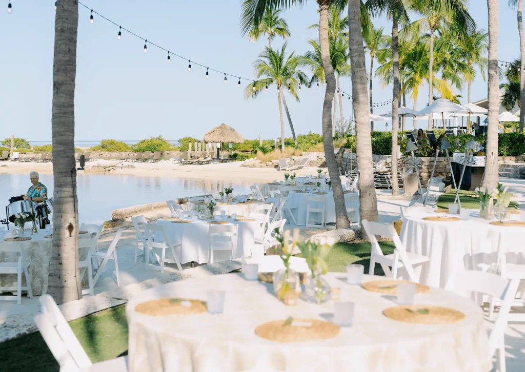 Outdoor event setup with round white tables and string lights on a sandy beach by a tropical lagoon.