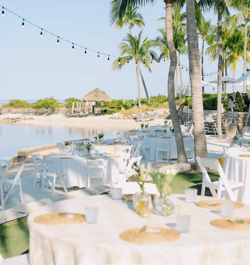 Outdoor event setup with round white tables and string lights on a sandy beach by a tropical lagoon.