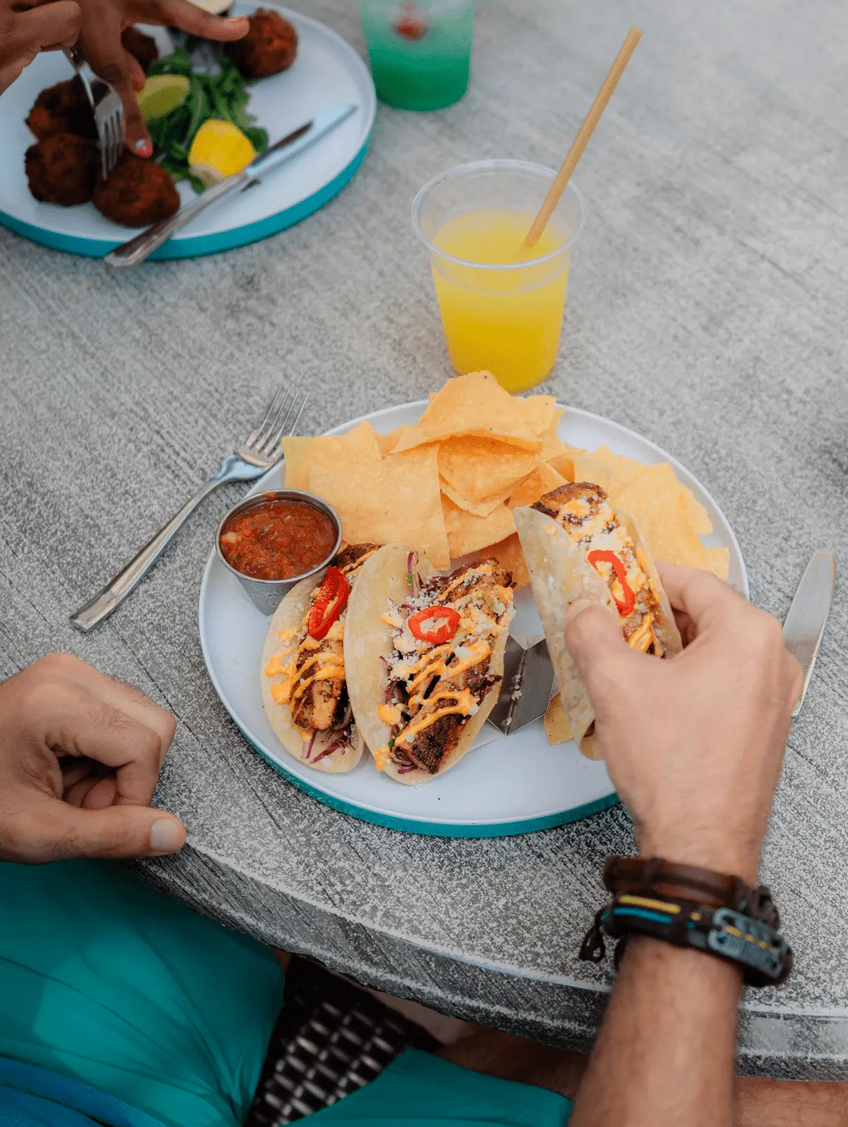 Close-up of fish tacos, tortilla chips, and salsa served with a bright yellow tropical drink outdoors.