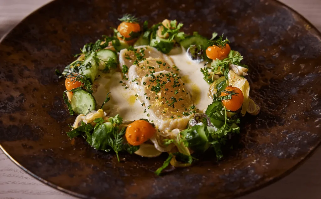Plated gourmet fish fillet with fresh herbs, vegetables, and creamy sauce arranged artfully on a dark ceramic plate.