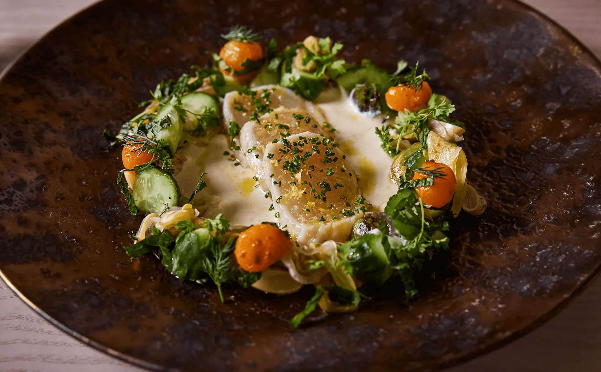 Gourmet crudo dish with thin slices of white fish, fresh herbs, cucumbers, and cherry tomatoes in a creamy sauce.