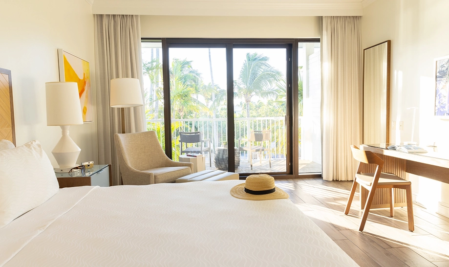 Sun-drenched hotel room with a white bed, straw hat, and sliding glass doors opening to a tropical balcony.