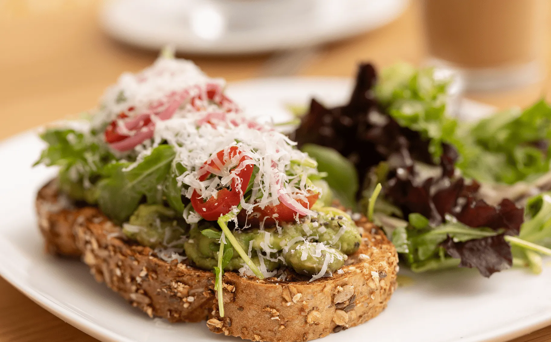 Avocado toast on multigrain bread topped with shaved cheese, tomatoes, and arugula served with a side salad.