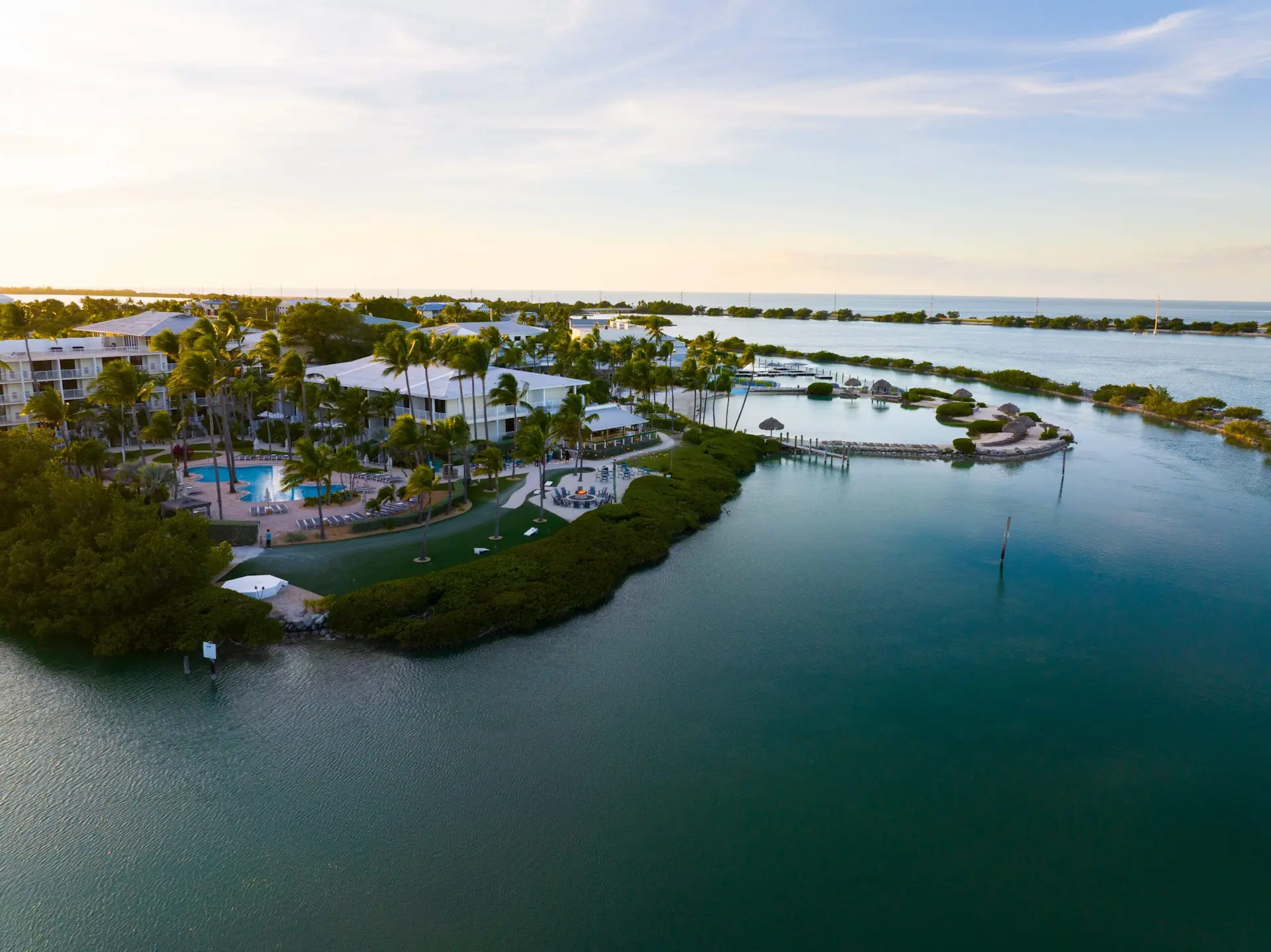 Aerial view of a tropical waterfront resort with palm trees, pools, docks, and calm blue waters at sunset.
