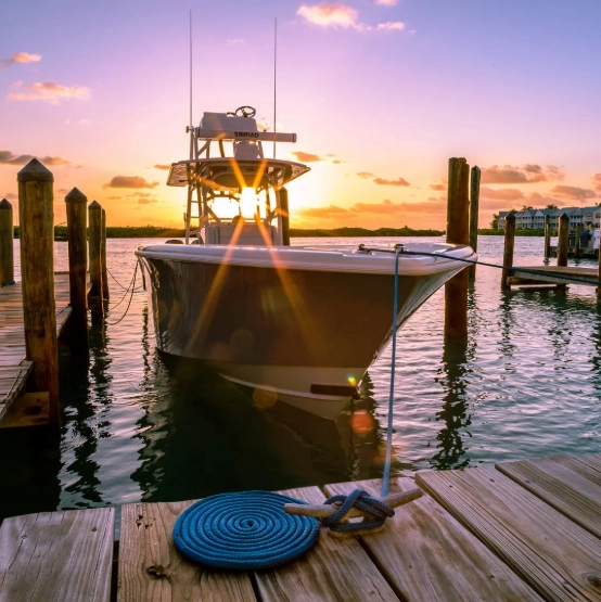 A fishing boat docked at a marina pier with a bright sunburst and colorful orange sunset in the background.