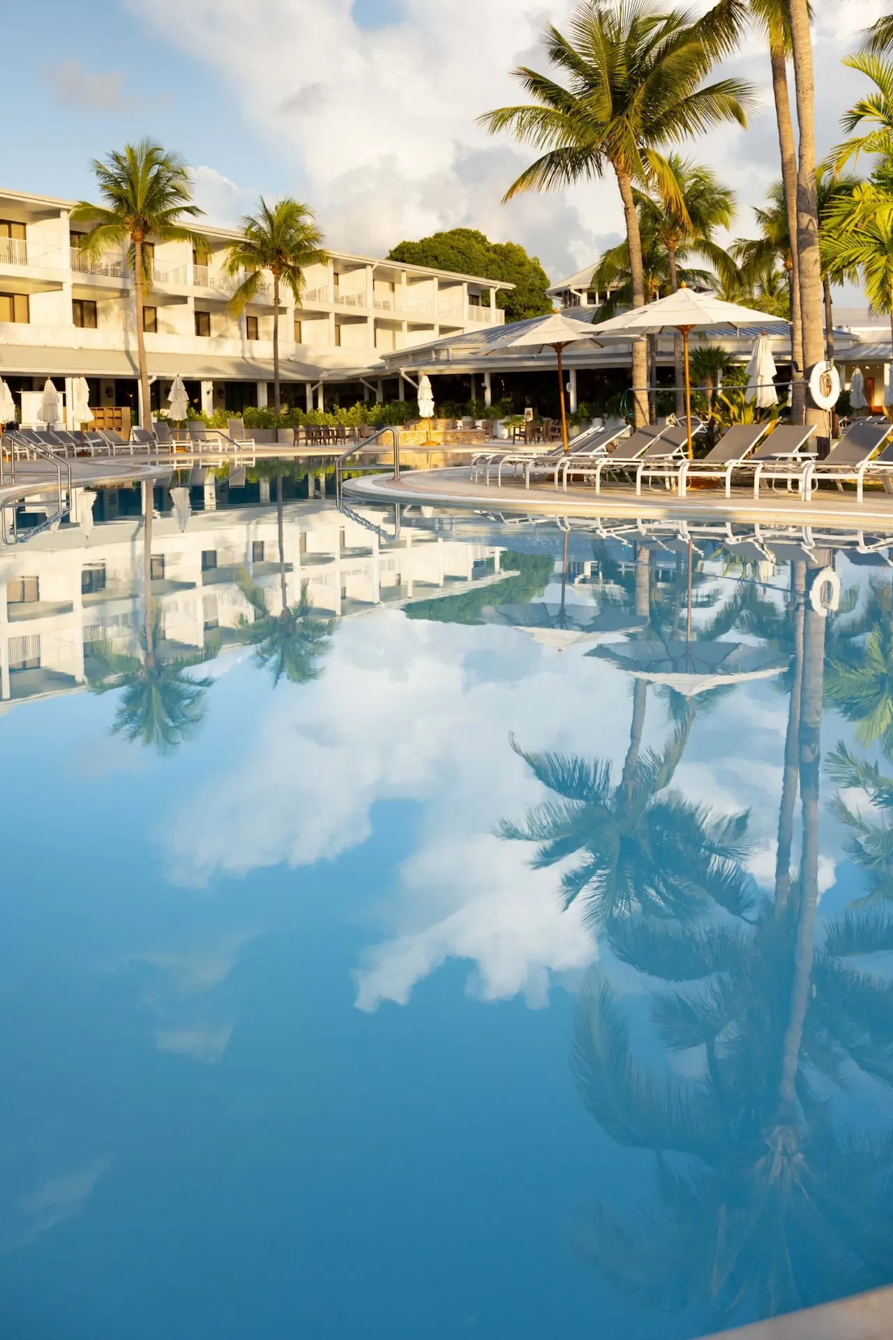 Clear resort swimming pool reflecting palm trees, white umbrellas, and the hotel building under a blue sky.