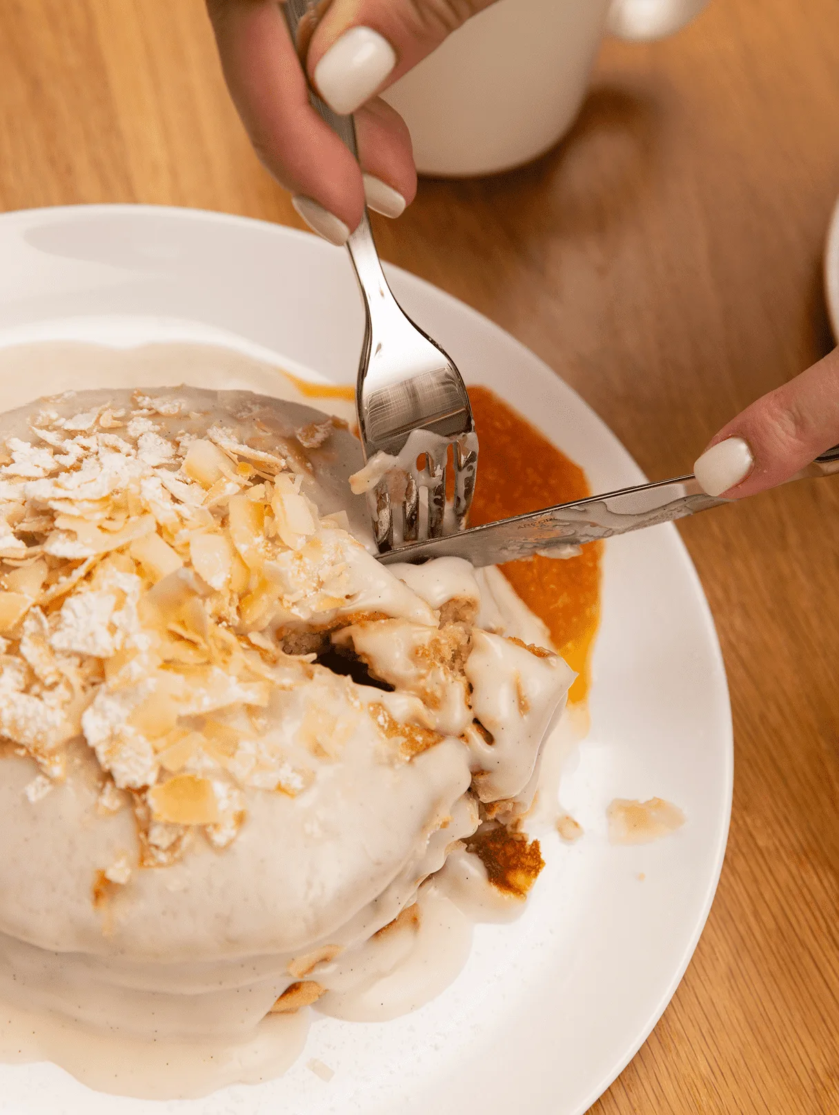 Close-up of hands using a fork and knife to cut into a stack of pancakes topped with white glaze and toasted coconut.