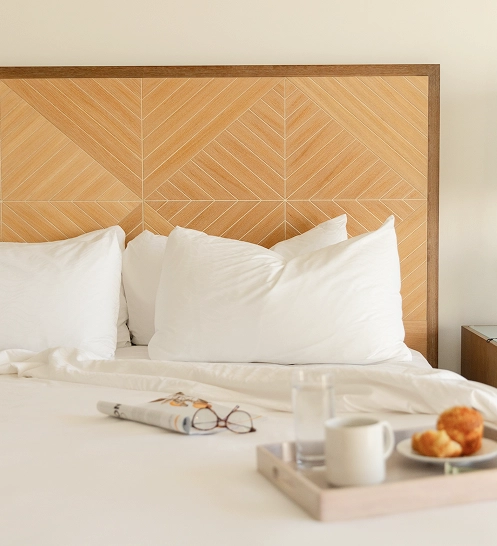 Minimalist bedroom with wooden headboard, white pillows, and breakfast tray with coffee and croissants on the bed.