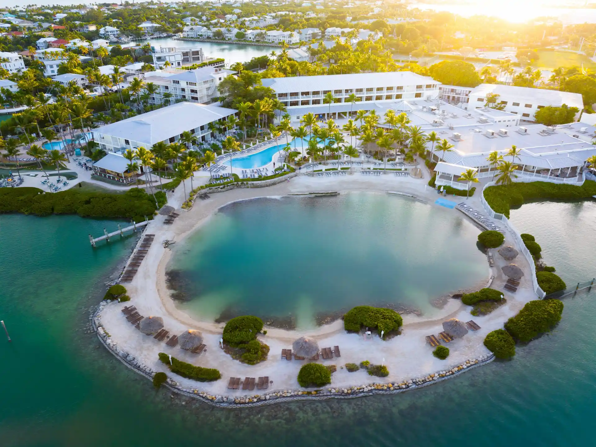 Aerial view of a tropical resort with lagoon-style pool, palm trees, waterfront beach area, and surrounding buildings at sunset.