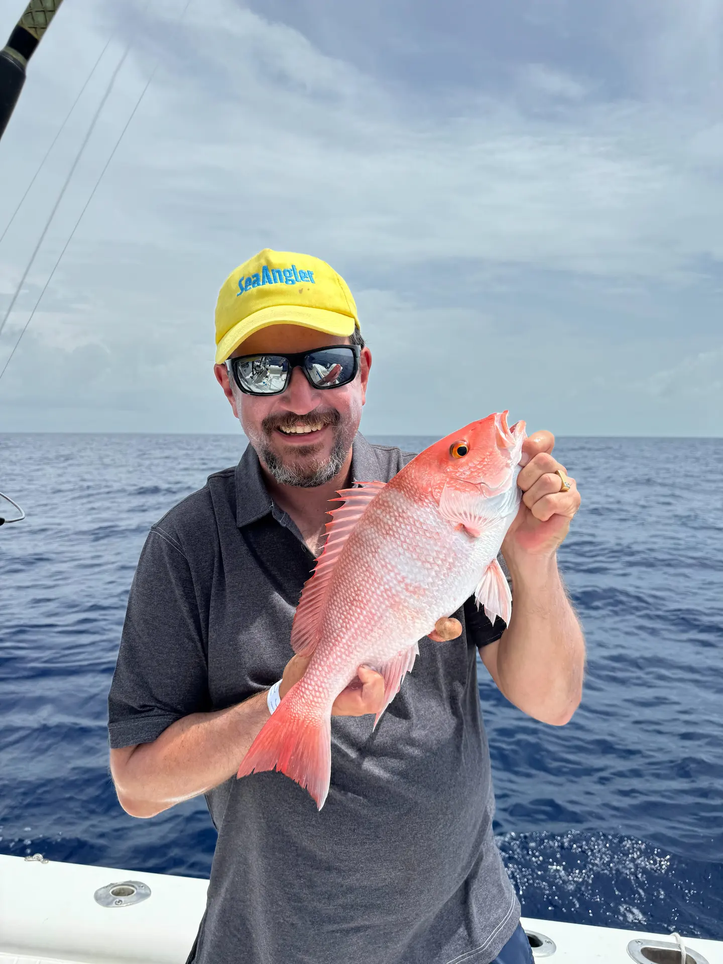 Man on a boat holding a red snapper fish, smiling, with open ocean in the background.