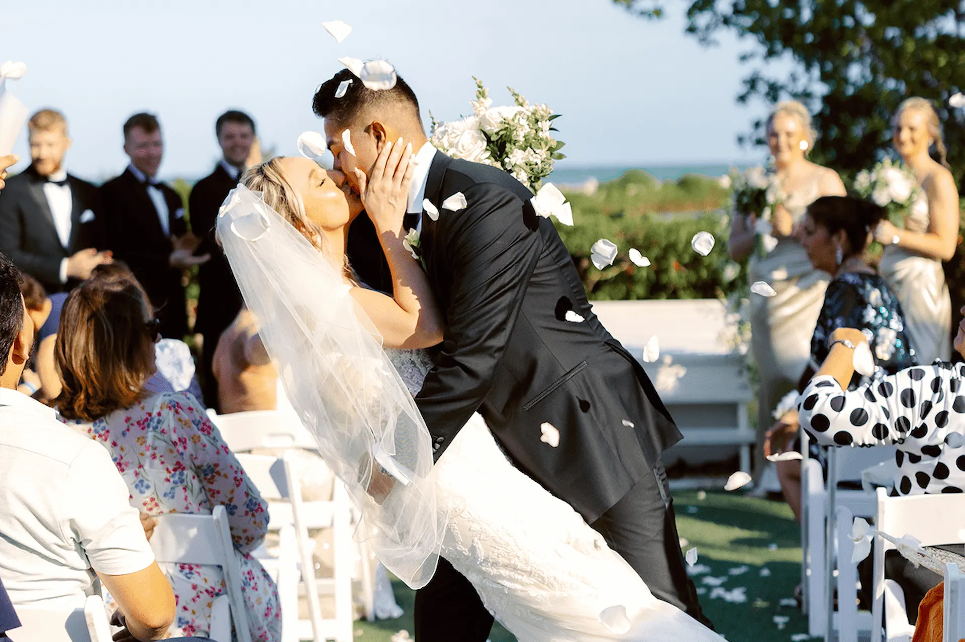 Bride and groom kissing during an outdoor wedding ceremony as guests toss white flower petals.
