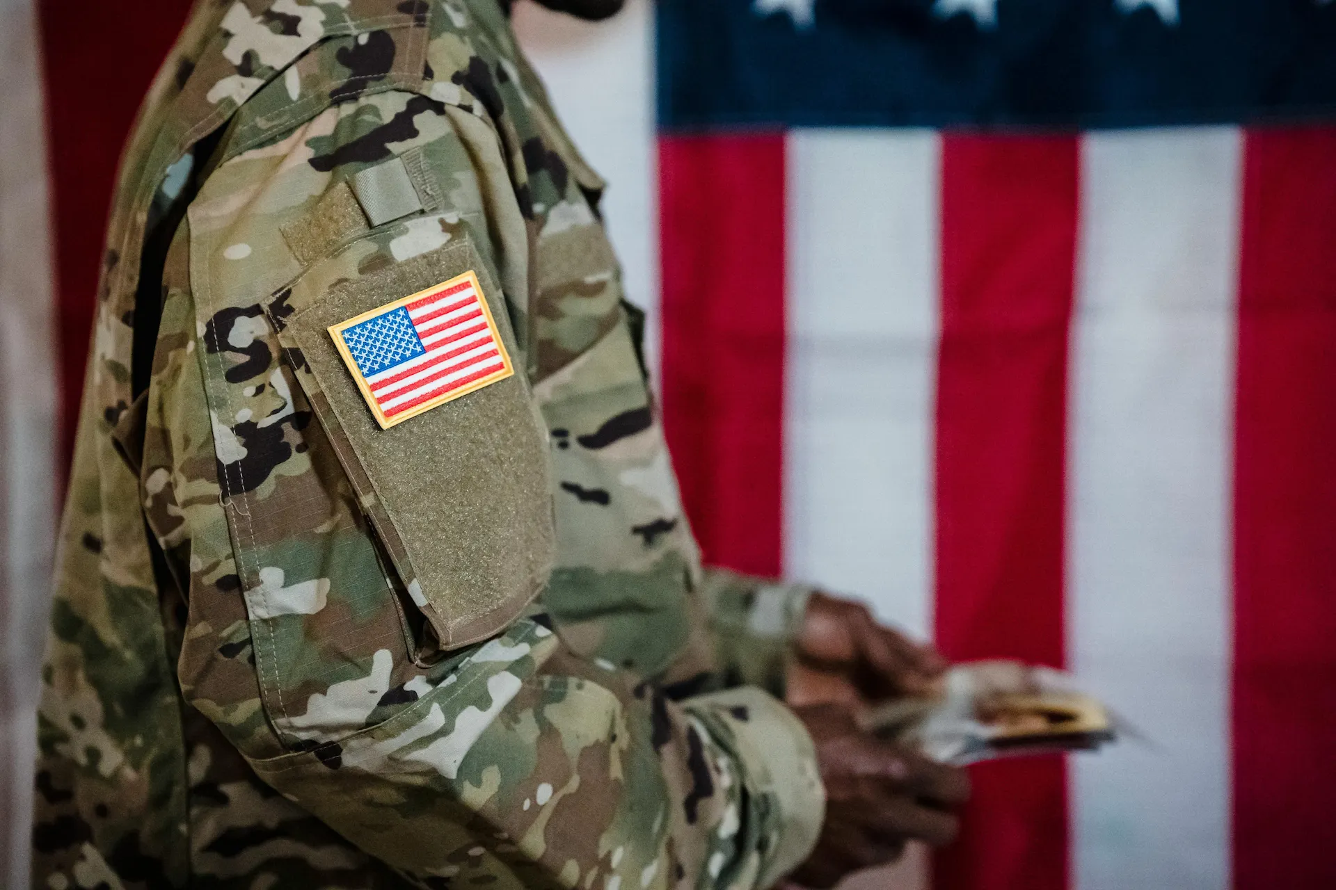 U.S. service member in camouflage uniform with an American flag patch, standing before a U.S. flag.