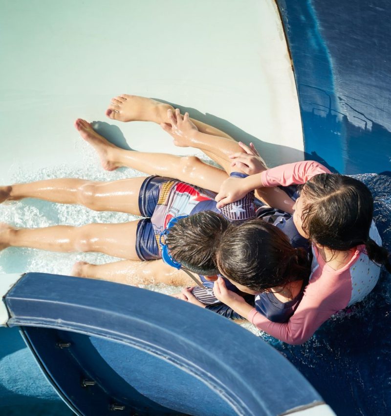 Three children race down a waterslide together at Coral Cay Adventure Park, splashing into the pool beneath the Florida Keys sun.